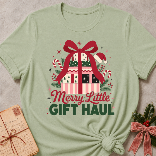 A sage green short-sleeve t-shirt with a festive graphic reading 'Merry Little GIFT HAUL' beneath a decorated gift box with a red bow. The shirt is styled flat on a textured gray background with a vintage notebook and holly accents.