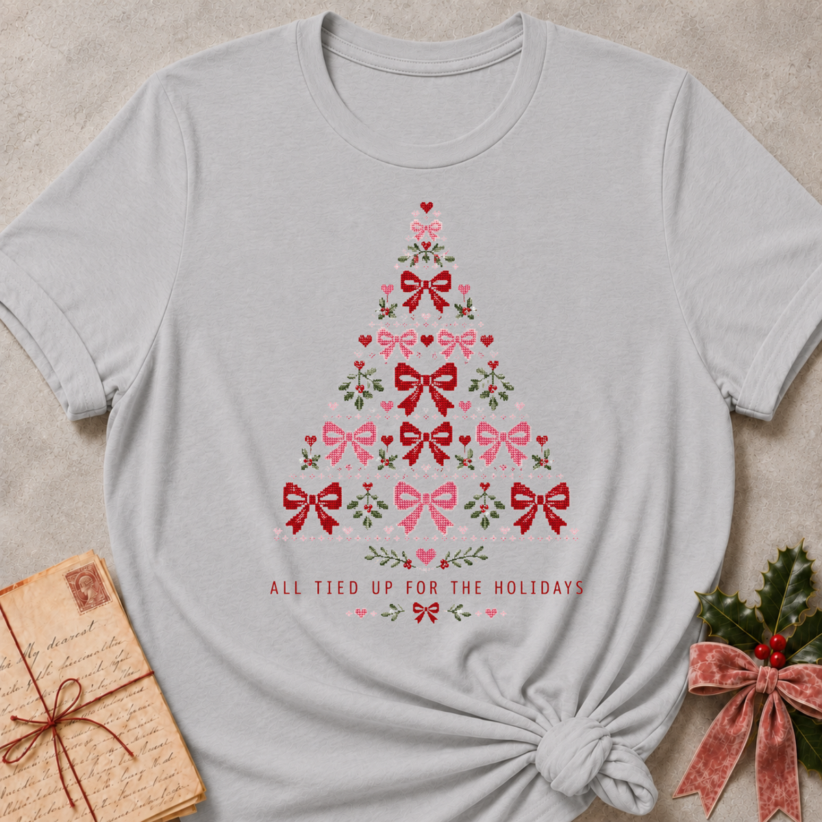 A light gray short-sleeve t-shirt with a Christmas tree made of red and pink bows and green leaves, featuring the text 'ALL TIED UP FOR THE HOLIDAYS' below. The shirt is styled flat on a textured beige background with a vintage notebook and holly in the corners.