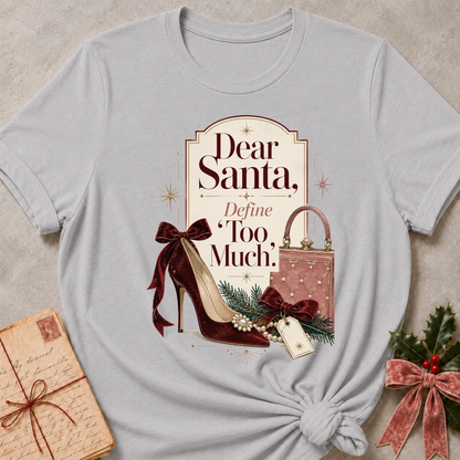 A light gray graphic t-shirt featuring a Christmas-themed design with the text 'Dear Santa, Define Too Much.' and illustrations of a burgundy high heel, a pink quilted handbag, and green pine branches, styled in a flat-lay with a vintage letter, gift box, and holly.