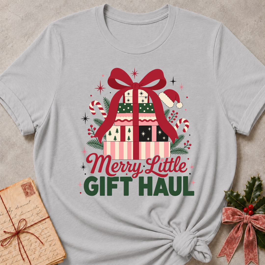 A light gray short-sleeve t-shirt with a festive Christmas graphic that reads 'Merry Little GIFT HAUL'. The graphic features a patterned gift box with a large red bow, candy canes, pine branches, and a Santa hat.