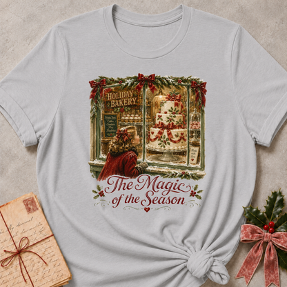 A light gray short-sleeve t-shirt with a vintage Christmas bakery graphic, styled flat on a textured beige background with a knotted hem. The graphic features a woman looking out of a bakery window at a decorated cake, with the text 'The Magic of the Season'.