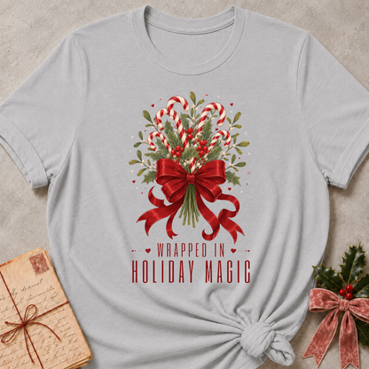 A light gray short-sleeve t-shirt with a festive Christmas graphic featuring candy canes, holly, red berries, a large red bow, and the text 'WRAPPED IN HOLIDAY MAGIC'. The shirt is styled flat on a textured beige background with a vintage postcard and a small holly sprig.