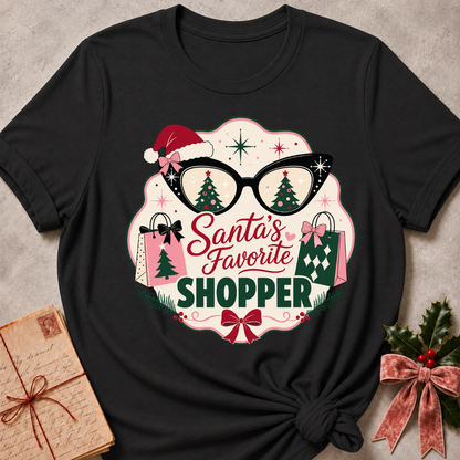 A black graphic t-shirt featuring a festive Christmas design with the text 'Santa's Favorite SHOPPER' in red and green, accompanied by glasses with Christmas tree reflections, shopping bags, and a Santa hat, presented in a styled flat-lay with holiday props.