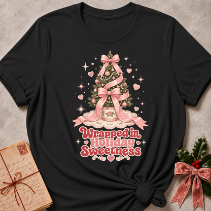 A black short-sleeve t-shirt with a pink Christmas tree graphic and the text 'Wrapped in Holiday Sweetness' printed on the front, styled in a flat-lay with a vintage letter and holly on a textured gray background.