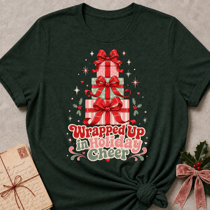A dark forest green t-shirt with a festive Christmas graphic of stacked gift boxes and the text 'Wrapped up in Holiday cheer'. The t-shirt is styled in a flat-lay with a vintage letter on the left and holly on the right, set against a neutral gray background.