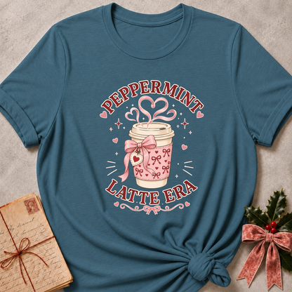 A teal blue graphic t-shirt featuring a pink and white coffee cup design with the text 'PEPPERMINT LATTE ERA'. The t-shirt is styled in a flat-lay arrangement on a textured gray background, accompanied by a vintage-style notebook and a small holly sprig with a pink ribbon.