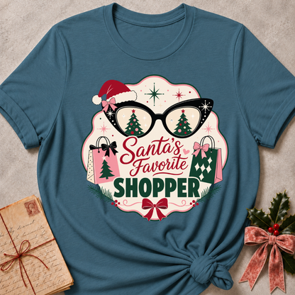 A teal blue graphic t-shirt featuring a festive Christmas design with the text 'Santa's Favorite SHOPPER' in red and green lettering, surrounded by glasses with Christmas tree reflections, shopping bags, a Santa hat, and bows. The t-shirt is styled flat on a neutral textured background with a vintage notebook and holly ribbon props.