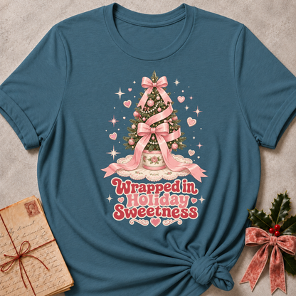 A teal blue short-sleeve t-shirt with a Christmas tree graphic and the text 'Wrapped in Holiday Sweetness' in pink and white. The t-shirt is styled flat on a textured gray background with a vintage notebook on the left and a holly sprig with a pink ribbon on the right.