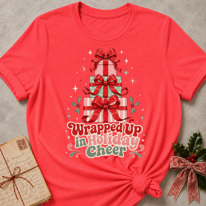 A heather Red short-sleeve t-shirt with a Christmas graphic of stacked presents and the text 'Wrapped Up in Holiday Cheer'. The shirt is styled in a flat-lay with a vintage notebook on the left and holly with a pink ribbon on the right, set against a textured gray background.