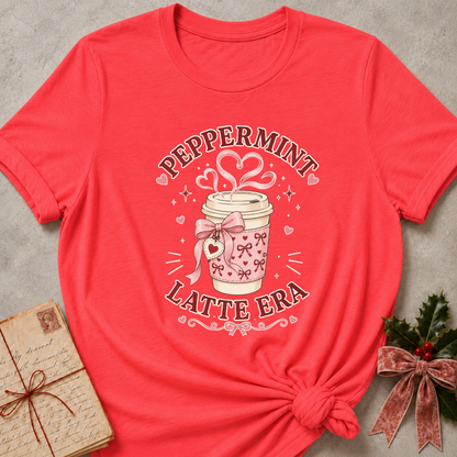 A coral pink short-sleeve graphic T-shirt laid flat on a light gray textured surface, styled with a vintage letter and a holly bow. The shirt features a central graphic of a pink patterned coffee cup with hearts and the text 'PEPPERMINT LATTE ERA'.