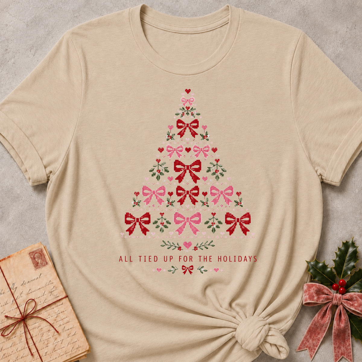 A beige short-sleeve t-shirt with a Christmas tree graphic made of red and pink bows, green holly leaves, and hearts, with the text 'ALL TIED UP FOR THE HOLIDAYS' below. The shirt is styled flat on a gray textured background with a vintage letter and a velvet ribbon bow.