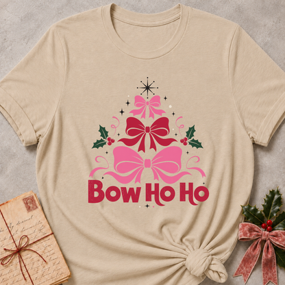 A beige short-sleeve t-shirt with a 'Bow Ho Ho' Christmas graphic, styled in a flat-lay with a vintage letter, greenery, and a velvet ribbon bow on a textured gray background.