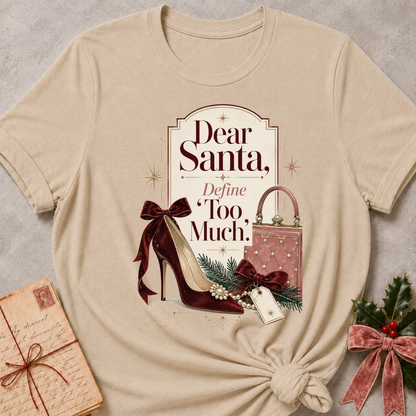 A beige graphic t-shirt with a Christmas-themed design reading 'Dear Santa, Define Too Much.' featuring a burgundy high heel, handbag, and pine branches, styled flat-lay with a vintage letter and holly on a gray textured background.