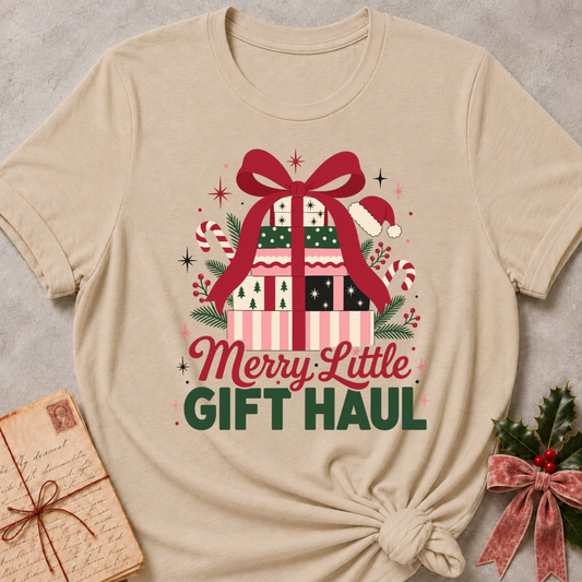 A beige short-sleeve T-shirt laid flat on a light gray textured surface, styled with a holiday gift graphic and the text 'Merry Little GIFT HAUL.' The shirt has a large Christmas present illustration with a red bow, green and pink striped pattern, candy canes, pine branches, and dark green lettering.