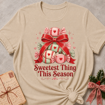 A beige short-sleeve t-shirt with a Christmas-themed graphic of wrapped candies under a red bow and the text 'Sweetest Thing This Season'. The t-shirt is styled flat on a gray textured background with a vintage letter and holly in the corners.