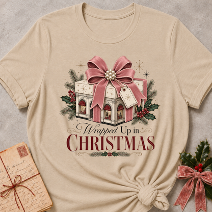 A beige short-sleeve t-shirt with a Christmas graphic print reading 'Wrapped Up in Christmas' under an illustration of a gift box with a pink bow, holly, and pine branches. The shirt is styled flat on a gray textured background with a vintage letter and greenery props.