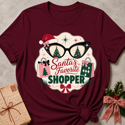 A maroon short-sleeve t-shirt with a festive graphic print reading 'Santa's Favorite SHOPPER' in red and green lettering, surrounded by glasses with Christmas trees, shopping bags, a Santa hat, and bows. The shirt is styled flat on a textured gray background with a vintage paper envelope and holly in the corners.