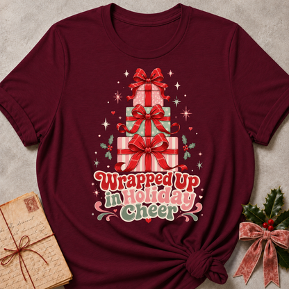 A maroon short-sleeve t-shirt with a Christmas graphic of stacked presents and the text 'Wrapped Up in Holiday Cheer'. The shirt is styled in a flat-lay with a vintage notebook on the left and holly with a pink ribbon on the right, set against a textured gray background.