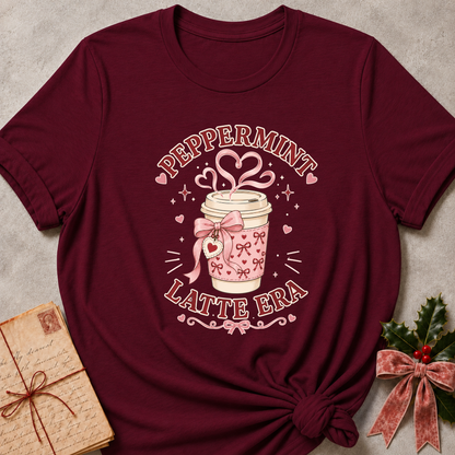 A maroon short-sleeve graphic T-shirt laid flat on a textured light gray surface, styled with a vintage notebook on the left and holly with a pink bow on the right. The shirt features a central graphic of a pink patterned coffee cup with a pink bow, hearts, and the words 'PEPPERMINT LATTE ERA'.