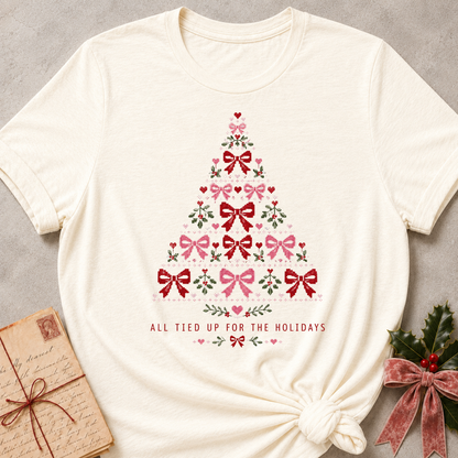 An off-white short-sleeve t-shirt with a Christmas tree graphic made of red and pink bows, green leaves, and hearts, with the text 'ALL TIED UP FOR THE HOLIDAYS' printed below. The t-shirt is styled flat on a gray textured background with a vintage book and holly in the corners.