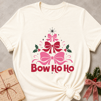 An off-white short-sleeve t-shirt with a 'Bow Ho Ho' Christmas graphic, styled in a flat-lay with a vintage letter and greenery on a gray textured background.