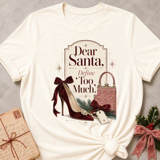 An off-white short-sleeve t-shirt with a Christmas-themed graphic print that reads 'Dear Santa, Define Too Much.' The print features a burgundy high heel, a pink patterned handbag, green pine branches, and red bows.
