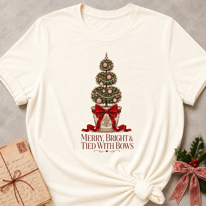 An off-white short-sleeve t-shirt with a Christmas tree graphic and the text 'MERRY, BRIGHT & TIED WITH BOWS' printed on the front, styled flat-lay on a gray textured background with a vintage letter and holly accents.