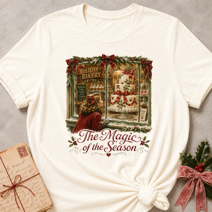 An off-white short-sleeve t-shirt with a Christmas-themed graphic print showing a woman looking out of a bakery window at a decorated cake, with the text 'The Magic of the Season'. The t-shirt is styled flat on a gray textured background with a vintage letter and holly accents.