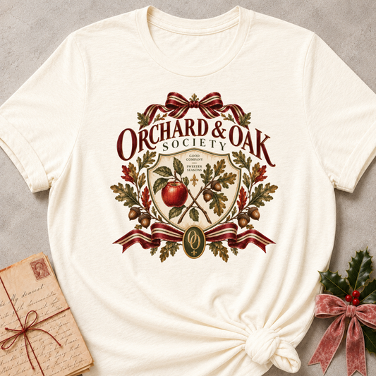 Natural cream women's relaxed tee with a vintage Orchard & Oak Society crest featuring a red apple, oak leaves, acorns, and striped ribbon bows