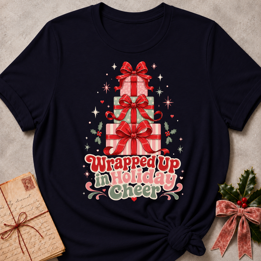 A navy blue short-sleeve t-shirt with a Christmas graphic of stacked gift boxes and the text 'Wrapped up in Holiday cheer'. The shirt is styled in a flat-lay on a textured gray background with a vintage notebook and holly in the corners.