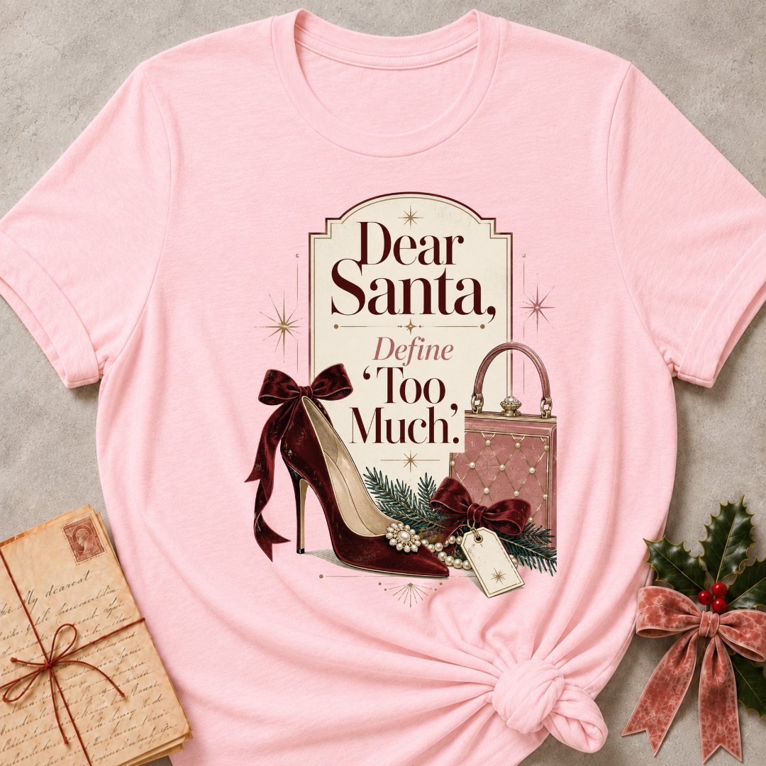 A light pink graphic t-shirt styled in a flat-lay arrangement on a textured gray background, featuring a vintage Christmas-themed print with the text 'Dear Santa, Define Too Much.' and illustrations of a high-heeled shoe, a handbag, and evergreen branches.