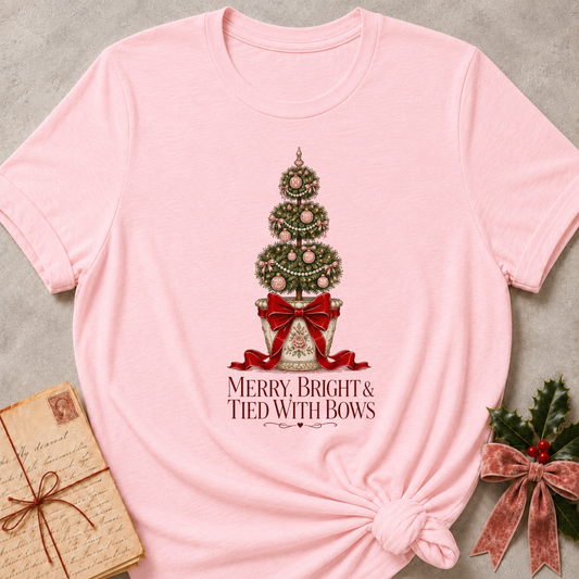 A light pink short-sleeve t-shirt with a Christmas tree graphic and the text 'MERRY, BRIGHT & TIED WITH BOWS' printed on the front. The t-shirt is styled in a flat-lay arrangement with a vintage letter, a holly sprig, and a pink ribbon on a textured gray background.