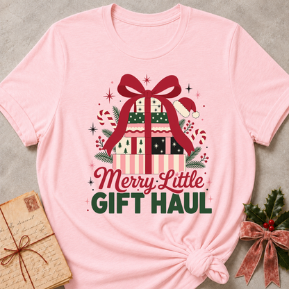 A light pink short-sleeve t-shirt with a 'Merry Little Gift Haul' graphic print, styled in a flat-lay with a vintage envelope and holly on a gray textured background.