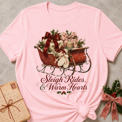A light pink short-sleeve t-shirt with a Christmas-themed graphic of a sleigh filled with gifts and holly, with the text 'Sleigh Rides & Warm Hearts' below it. The shirt is styled flat on a gray textured background with a vintage envelope and holly sprig as props.