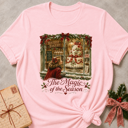 A light pink short-sleeve t-shirt with a Christmas-themed graphic print of a woman looking out of a bakery window at a decorated cake, with the text 'The Magic of the Season'. The shirt is styled flat on a gray textured background with a vintage envelope and holly accents.