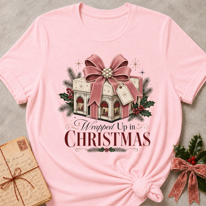 A light pink short-sleeve t-shirt with a Christmas-themed graphic print. The graphic features an illustration of a wrapped gift box under a large dusty rose bow, surrounded by pine branches, holly leaves, and a tag, with the text 'Wrapped Up in CHRISTMAS' below.