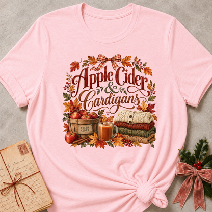 Pink relaxed fit tee with Apple Cider and Cardigans lettering, a cider mug, a basket of red apples, stacked sweaters and a gingham bow