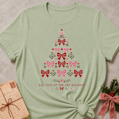 A sage green short-sleeve t-shirt with a Christmas tree made of pink and red bows and floral details, styled flat-lay on a textured gray background with a vintage book and holly in the corners.