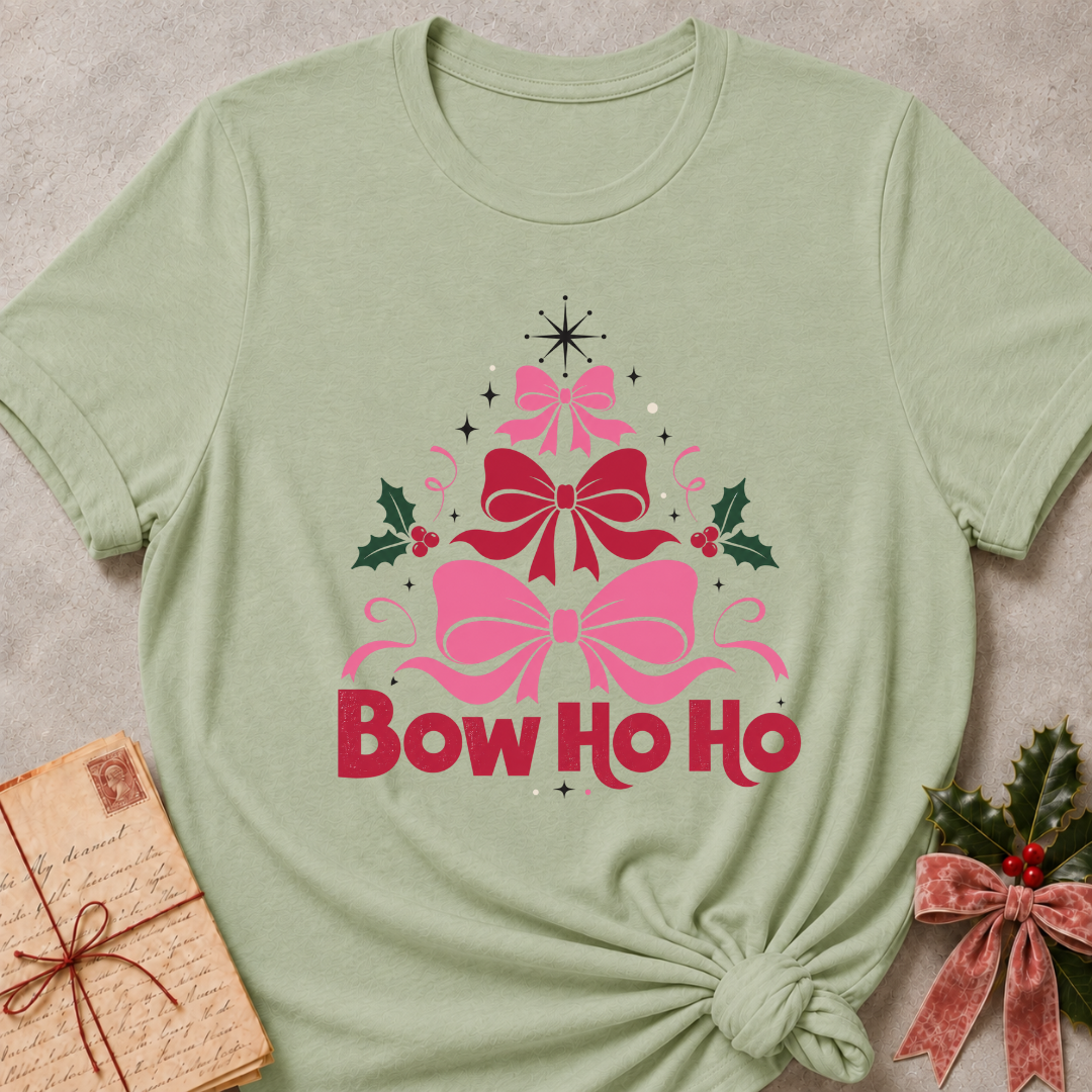 A sage green short-sleeve t-shirt with a 'Bow Ho Ho' Christmas graphic, styled in a flat-lay with a vintage letter and a velvet bow on a neutral textured background.