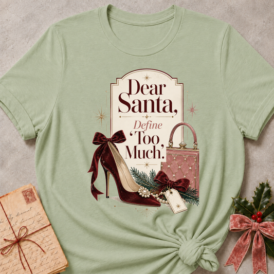 A sage green graphic t-shirt styled in a flat-lay with a knotted hem, featuring a vintage-inspired Christmas graphic with the text 'Dear Santa, Define Too Much.' and illustrations of a burgundy high heel, a pink purse, and green pine branches.
