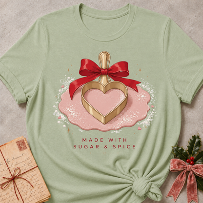 A sage green short-sleeve t-shirt with a festive graphic of a gold heart-shaped cookie cutter in a pink bowl tied with a red ribbon, with the text 'MADE WITH SUGAR & SPICE' below. The t-shirt is styled flat on a textured gray background with a vintage letter and holly accents.