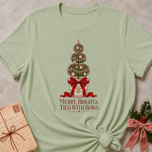 A sage green short-sleeve t-shirt with a Christmas tree graphic and the text 'MERRY, BRIGHT & TIED WITH BOWS' printed on the front, styled in a flat-lay with a vintage notebook and holly on a textured gray background.