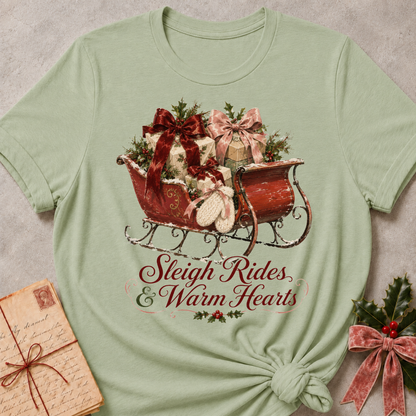 A sage green short-sleeve t-shirt with a Christmas-themed graphic of a red sleigh filled with gifts and holly, with the text 'Sleigh Rides & Warm Hearts' printed below. The t-shirt is styled flat on a textured gray background with a vintage letter and holiday greenery props.