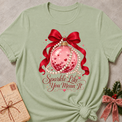A sage green short-sleeve t-shirt with a festive Christmas graphic, styled in a flat-lay with a vintage letter and holly. The graphic features a pink glitter ornament, red ribbon, white pearls, and the text 'Sparkle Like You Mean It'.
