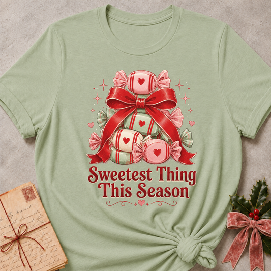 A sage green short-sleeve t-shirt with a Christmas-themed graphic of red and pink wrapped candies under a large red bow, with the text 'Sweetest Thing This Season' printed below. The shirt is styled flat on a textured gray background with a vintage letter and holly accents.