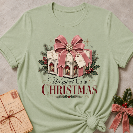 A sage green short-sleeve t-shirt with a Christmas graphic print reading 'Wrapped Up in CHRISTMAS' beneath an illustration of a gift box with a large pink bow, evergreen branches, and holly. The shirt is styled flat on a textured gray background with a vintage lettered envelope and holly in the corners.