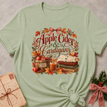 Sage relaxed fit tee with Apple Cider and Cardigans lettering, a cider mug, a basket of red apples, stacked sweaters and a gingham bow