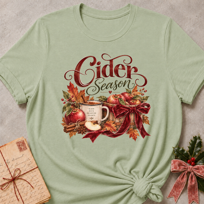 Sage women's relaxed tee with a Cider Season script design with a cider mug, red apples, cinnamon sticks, and a burgundy ribbon bow