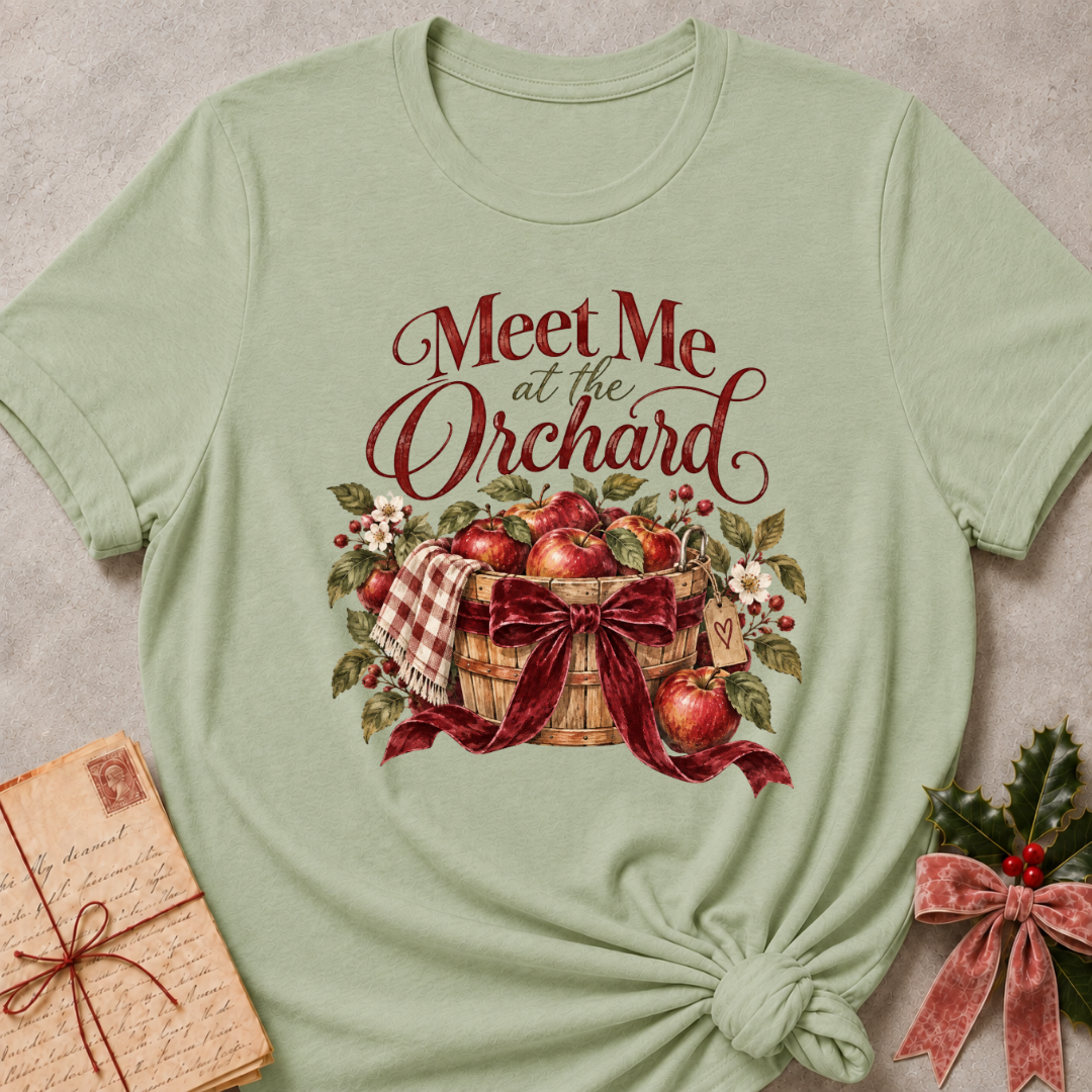 Sage relaxed fit tee with Meet Me at the Orchard lettering, a harvest basket of red apples, blossoms and a burgundy bow