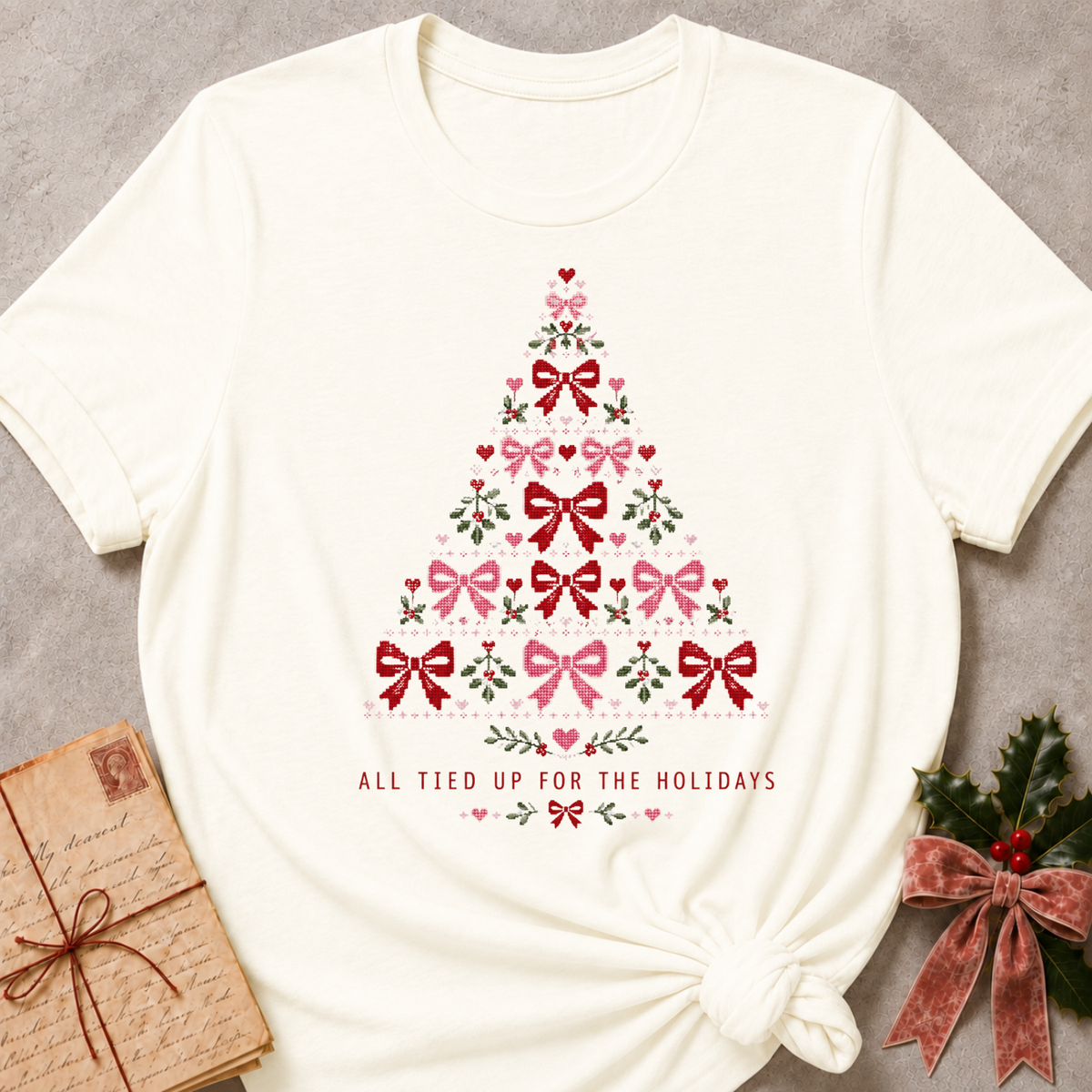 An off-white short-sleeve t-shirt with a Christmas tree graphic made of red and pink bows, green leaves, and hearts, with the text 'ALL TIED UP FOR THE HOLIDAYS' printed below. The t-shirt is styled in a flat-lay with a vintage lettered gift box on the left and holly with a red bow on the right, set against a textured gray background.
